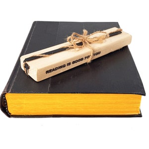 May include: A black book with yellow-edged pages, topped with a wooden pen holder tied with twine. The pen holder has the text "READING IS GOOD FOR YOU" printed on it. The book and pen holder are set against a white background.