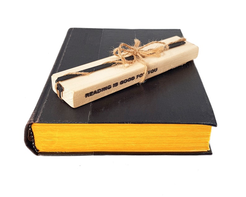 May include: A black book with yellow-edged pages, topped with a wooden pen holder tied with twine. The pen holder has the text "READING IS GOOD FOR YOU" printed on it. The book and pen holder are set against a white background.