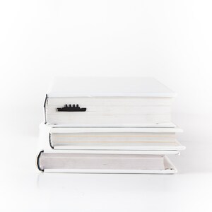 May include: A stack of three white hardcover books with a black ship bookmark on the top book.