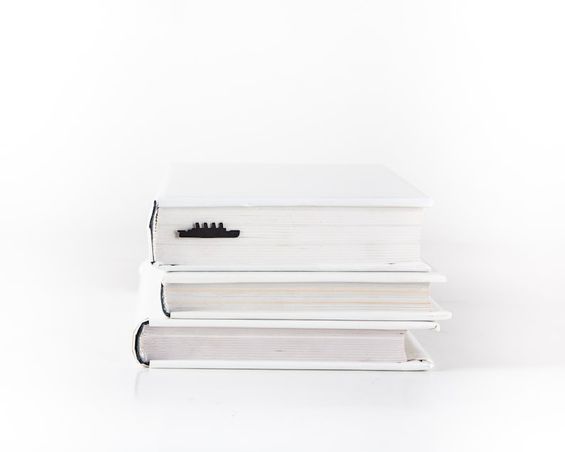 May include: A stack of three white hardcover books with a black ship bookmark on the top book.