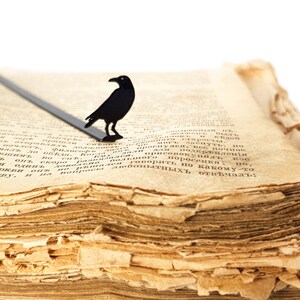 Black metal raven bookmark resting on the yellowed pages of an old book. A gothic, Poe-inspired page saver for readers who love mystery and the macabre.