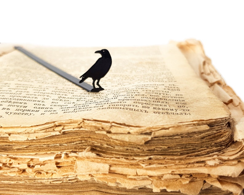 Black metal raven bookmark resting on the yellowed pages of an old book. A gothic, Poe-inspired page saver for readers who love mystery and the macabre.