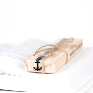 May include: A wooden bookmark with a metal anchor charm tied with twine. The bookmark is resting on a white book.