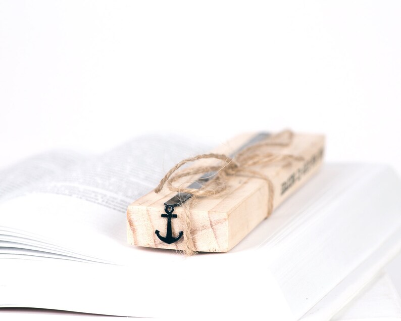 May include: A wooden bookmark with a metal anchor charm tied with twine. The bookmark is resting on a white book.