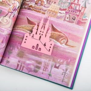 A pink metal castle shaped bookmark with a detailed design. The bookmark is placed on an open book with a colorful illustration of a castle and a fish.