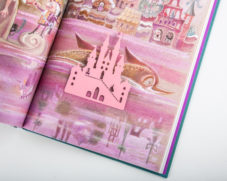 A pink metal castle shaped bookmark with a detailed design. The bookmark is placed on an open book with a colorful illustration of a castle and a fish.