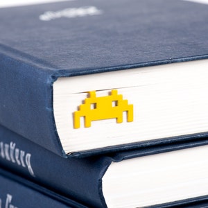 May include: A stack of three navy blue hardcover books with white pages. A yellow plastic bookmark shaped like a space invader is on the top book.