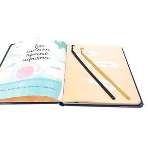 May include: Two gold and black metal bookmarks with an anchor design. The bookmarks are laying open on a page of a book with a blue cover. The page has a light brown background with a hand-drawn illustration of a sun and clouds. The text on the page reads "Tym можна просто мріяти" and "САЖАЙ МАЛЮЙ. ЗБЕРІГАЙ ФОТОГРАФІЇ. РОБИ КОЛАЖ".