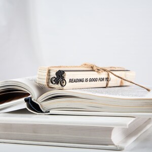 A stack of two books with a black meta bookmark Bike race on the wooden packaging block. The bookmark has a black silhouette of a person riding a bicycle and the text on the wooden block says 'READING IS GOOD FOR YOU'.