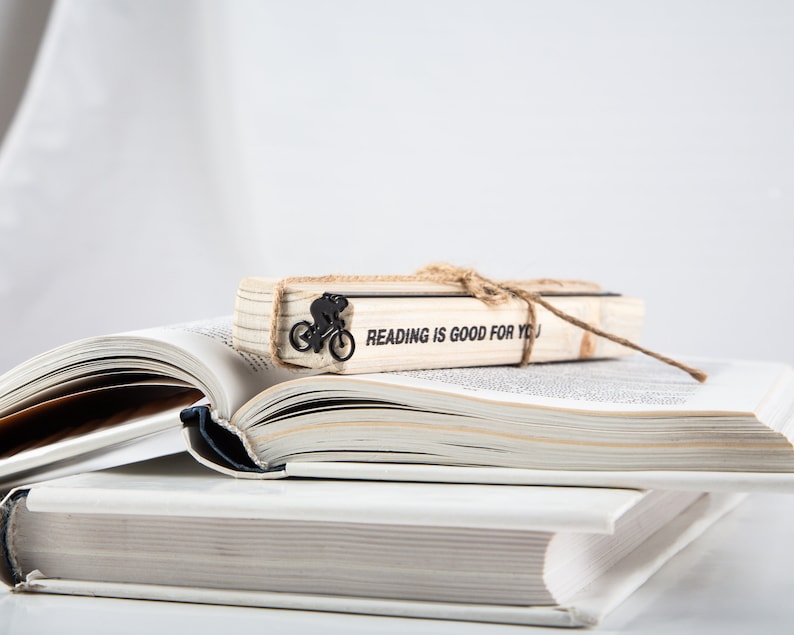 A stack of two books with a black meta bookmark Bike race on the wooden packaging block. The bookmark has a black silhouette of a person riding a bicycle and the text on the wooden block says 'READING IS GOOD FOR YOU'.
