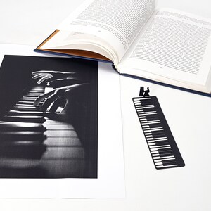 May include: A black and white print of piano keys with hands playing. A black metal bookmark shaped like piano keys with a silhouette of a person playing the piano. An open book with blue cover is also in the image.