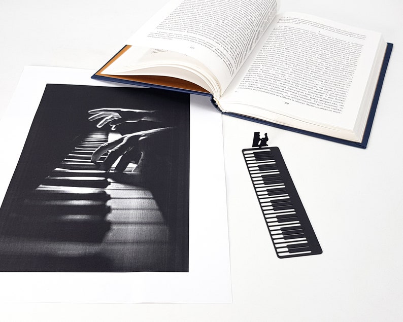 May include: A black and white print of piano keys with hands playing. A black metal bookmark shaped like piano keys with a silhouette of a person playing the piano. An open book with blue cover is also in the image.