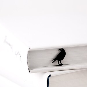 Black metal raven-shaped page saver perched on the edge of a white hardcover book. A dark, poetic gift for readers drawn to mystery and gothic themes.