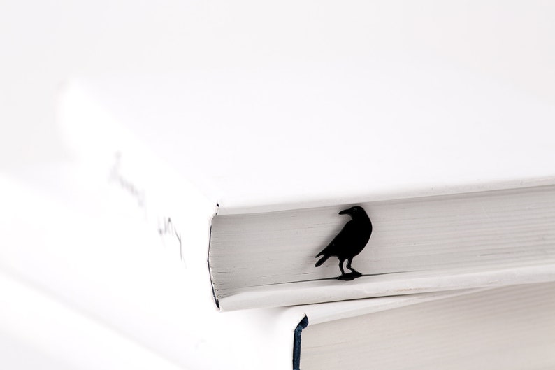 Black metal raven-shaped page saver perched on the edge of a white hardcover book. A dark, poetic gift for readers drawn to mystery and gothic themes.