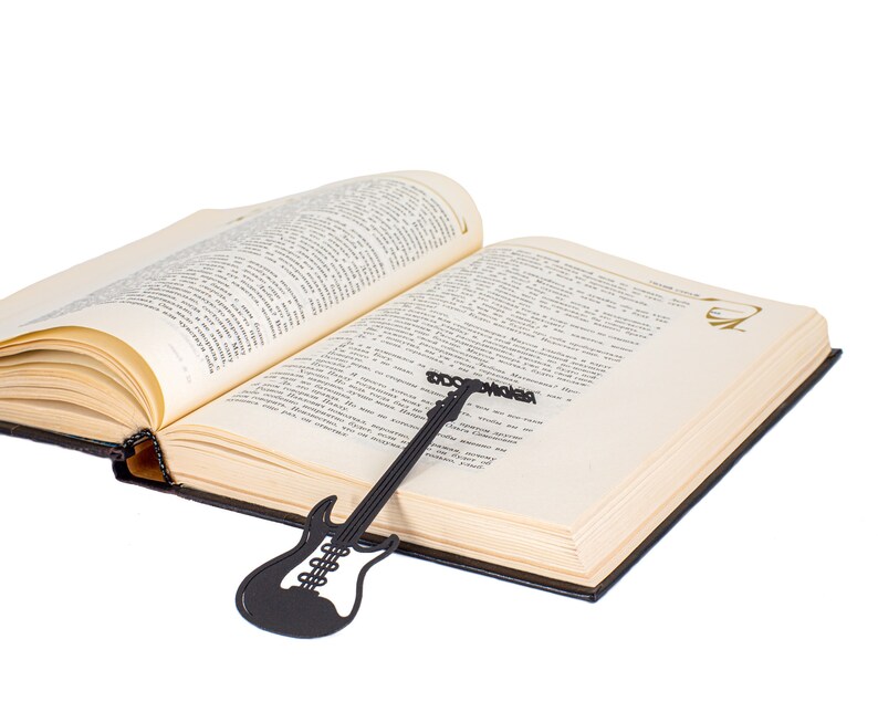 Black metal bookmark shaped like an electric guitar, with the words READING ROCKS running along the neck, placed inside an open book. A perfect gift for music lovers and bookworms.