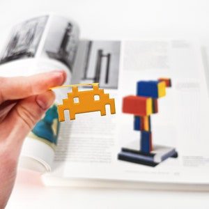May include: A yellow plastic space invader bookmark is held in a hand over an open book. The book has a colorful abstract painting on the right page.