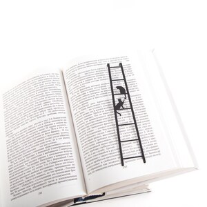 Black metal bookmark Cat on the Ladder. Two cats are sitting on the ladder, which is the size of an average book. The ladder stays between the pages of the book. The tips of the ladder are on the side of the book.