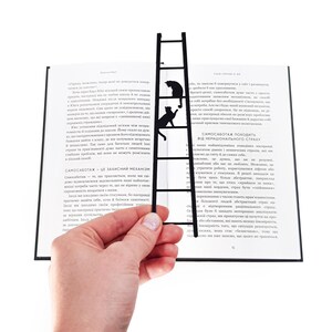 Hand holding a black metal cat bookmark shaped like a ladder with two cats climbing, positioned in front of an open book.