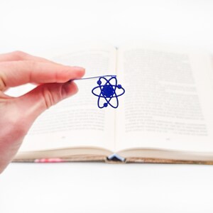 May include: A blue atom bookmark with a silver wire. The bookmark is held in a hand, with a book in the background.