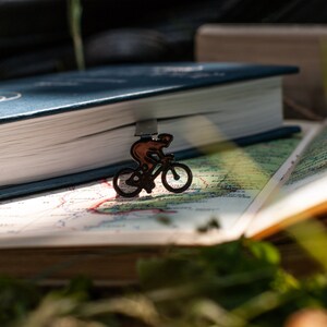 A black metal bookmark shaped like a cyclist riding a bicycle. The bookmark is on top of a closed blue book and an open book with a map printed on the pages.