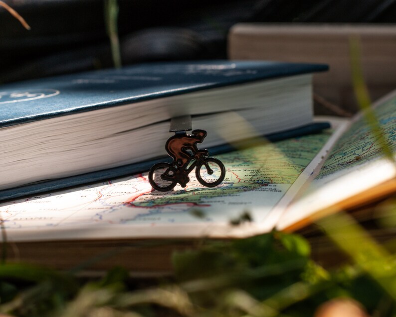 A black metal bookmark shaped like a cyclist riding a bicycle. The bookmark is on top of a closed blue book and an open book with a map printed on the pages.
