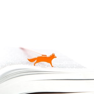 May include: An orange fox bookmark on a white book. The bookmark is made of metal and has a simple design.