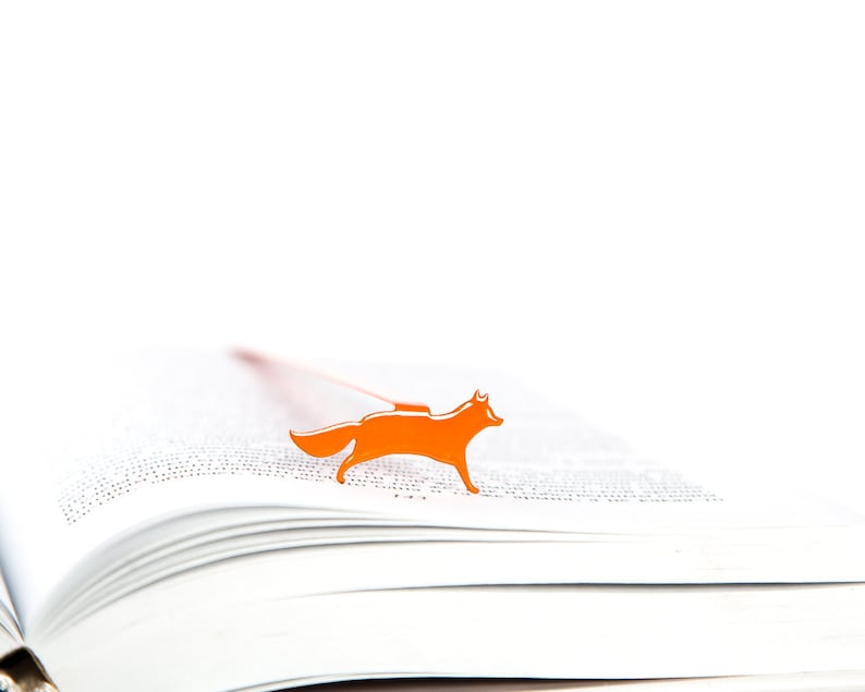 May include: An orange fox bookmark on a white book. The bookmark is made of metal and has a simple design.