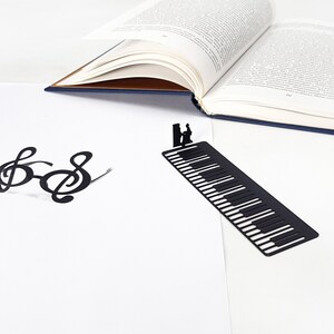 May include: A black metal bookmark shaped like a piano keyboard with a silhouette of a person playing the piano. A pair of black treble clef glasses and an open book are also in the image. The background is white.