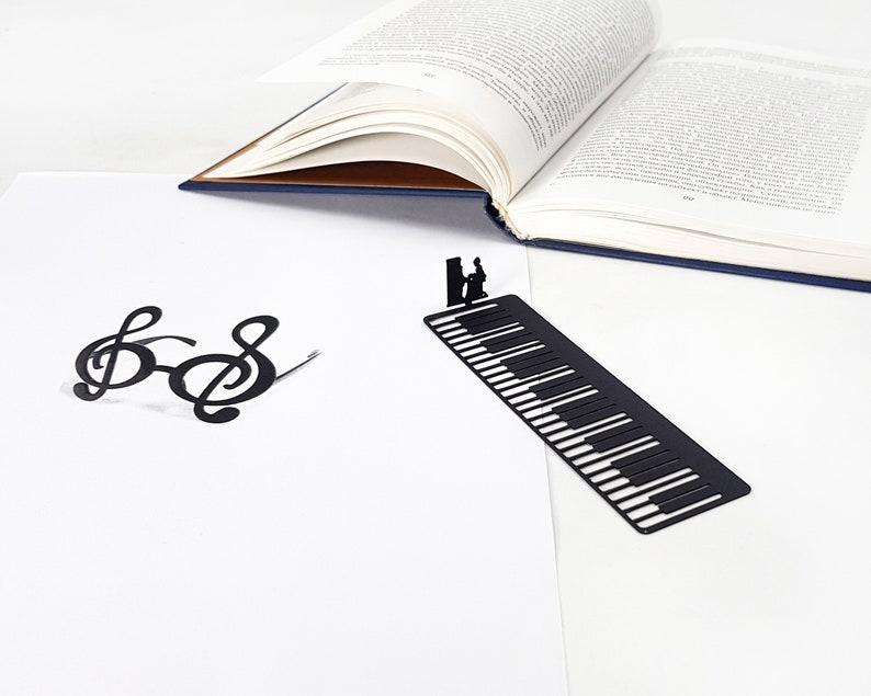 May include: A black metal bookmark shaped like a piano keyboard with a silhouette of a person playing the piano. A pair of black treble clef glasses and an open book are also in the image. The background is white.