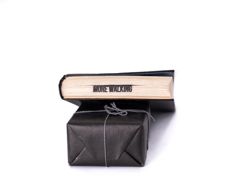 May include: A black book wrapped in black paper with a string bow. The book has the words "MORE WALKING" printed on the spine.