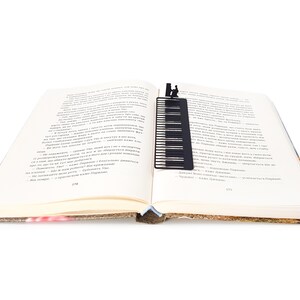 May include: A black metal piano key bookmark is inserted into an open book. The bookmark features a piano keyboard design with a silhouette of a person playing the piano at the top. The book's pages are cream-colored, and the spine is visible.