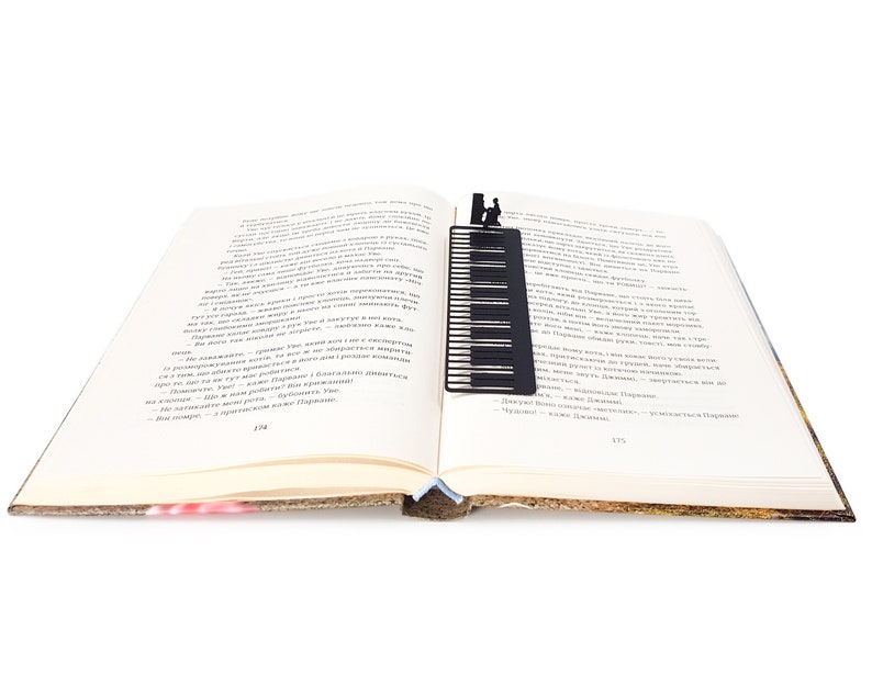 May include: A black metal piano key bookmark is inserted into an open book. The bookmark features a piano keyboard design with a silhouette of a person playing the piano at the top. The book's pages are cream-colored, and the spine is visible.