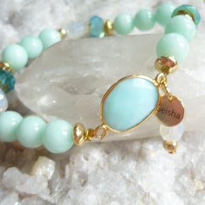 May include: A bracelet with pale green beads, gold details, and a large, light blue oval stone. A small gold charm with the word "Geisha" hangs from the oval stone. The bracelet is displayed on a white surface.