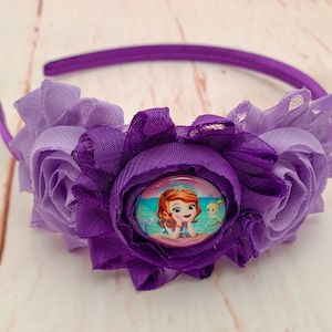 May include: Purple headband with two fabric flowers and a button featuring a cartoon princess with a mermaid tail.