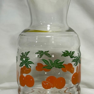 May include: A clear glass pitcher with a yellow lid and an orange and green citrus design. The pitcher is perfect for serving juice or water.