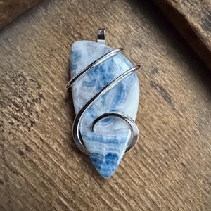 May include: A blue and white agate pendant wrapped in silver wire. The pendant is shaped like a teardrop and has a natural, organic look.