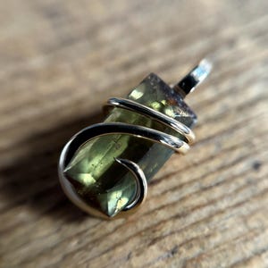 May include: A pendant featuring a rectangular, translucent olive-green gemstone wrapped in a polished gold-toned metal wire. The stone has a faceted surface and is secured by the wire, which forms a small loop at the top for attachment to a chain.