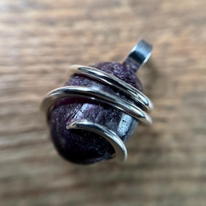 May include: A pendant featuring a smooth, dark purple, oval-shaped gemstone wrapped in polished silver-colored wire. The wire coils around the center of the stone, extending upward to form a small loop at the top for attachment to a chain or cord.