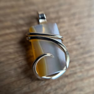 May include: A rectangular pendant with a silver bail and wire wrap. The stone is a mix of yellow, white, and gray. The wire wraps around the stone in a decorative pattern. The pendant is displayed on a wooden surface.
