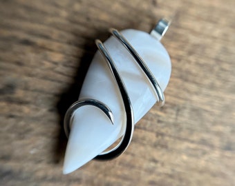 May include: A white teardrop-shaped pendant with a silver wire wrapped around it. The pendant is on a wooden surface.