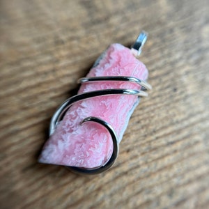 May include: A pink crystal pendant wrapped in silver wire. The crystal has a rough, textured surface and is a light pink color.