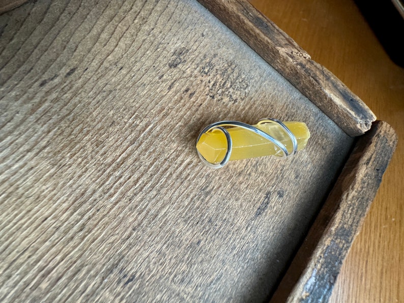Pu&ograve; includere: Un cristallo giallo avvolto da un anello di filo d'argento. Il cristallo &egrave; su una superficie di legno.