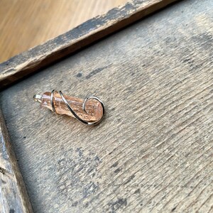 May include: A pink and orange crystal wrapped in silver wire, forming a pendant or charm.