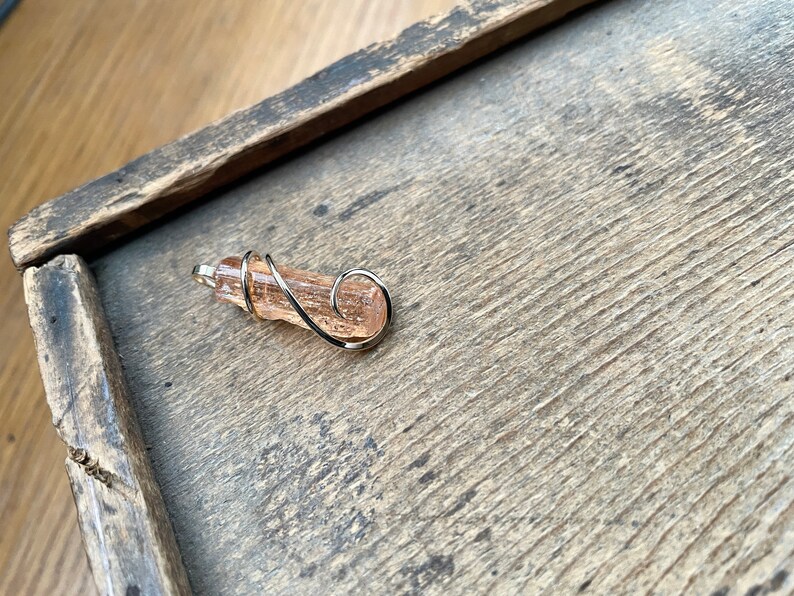 May include: A pink and orange crystal wrapped in silver wire, forming a pendant or charm.
