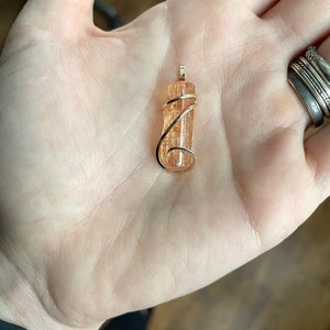 May include: A gold wire wrapped pendant featuring a natural orange and brown crystal. The crystal is a long, thin, rectangular shape.