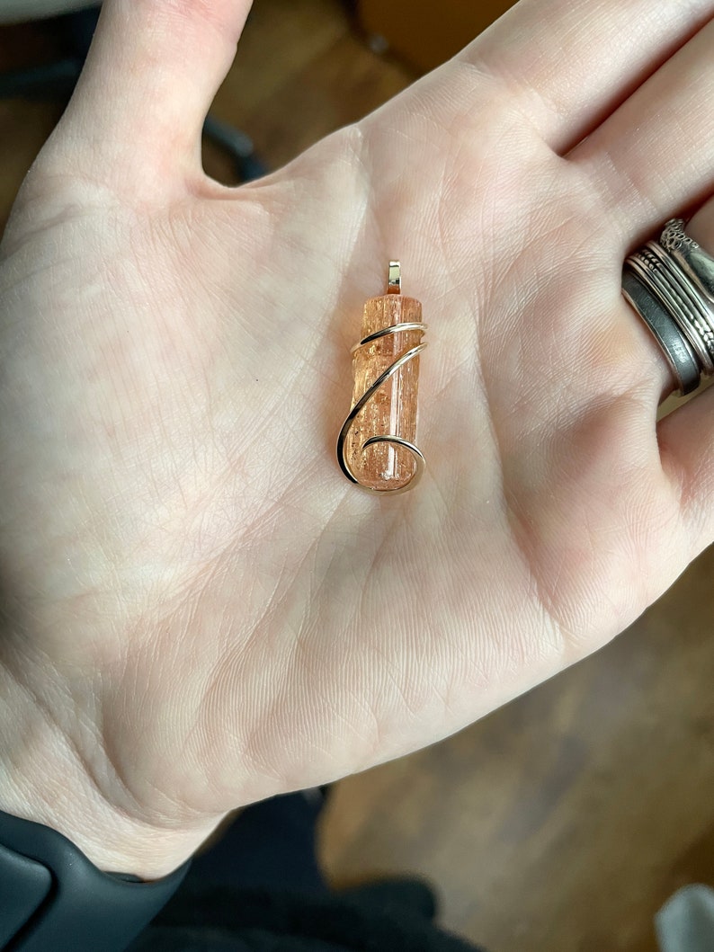 May include: A gold wire wrapped pendant featuring a natural orange and brown crystal. The crystal is a long, thin, rectangular shape.