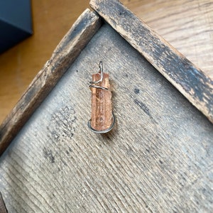 May include: A golden brown crystal pendant wrapped in silver wire. The pendant is sitting on a wooden surface.