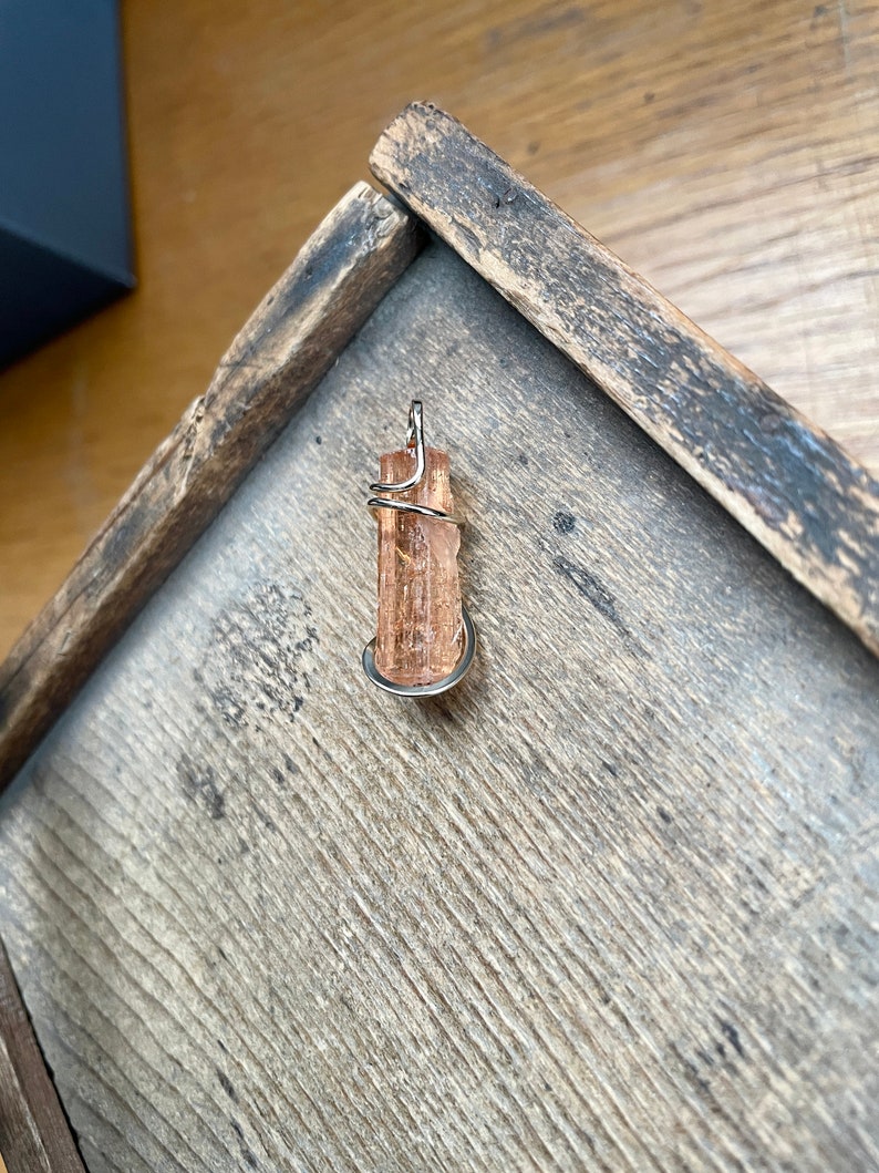 May include: A golden brown crystal pendant wrapped in silver wire. The pendant is sitting on a wooden surface.