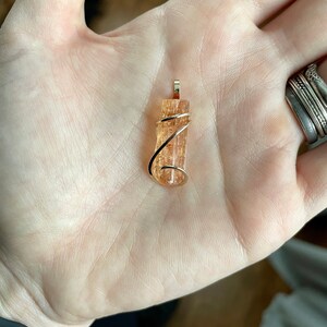 May include: A gold wire wrapped pendant featuring a natural, translucent, orange-brown crystal. The crystal has a striated texture and is approximately 1 inch in length.