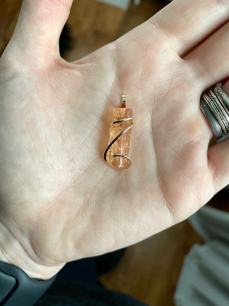 May include: A gold wire wrapped pendant featuring a natural, translucent, orange-brown crystal. The crystal has a striated texture and is approximately 1 inch in length.